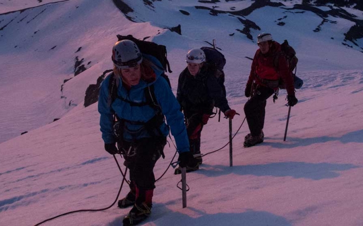 Three people wearing mountaineering gear are attached to each other by a rope. The snow appears pink and purple, presumably from the sunset or sunrise. 